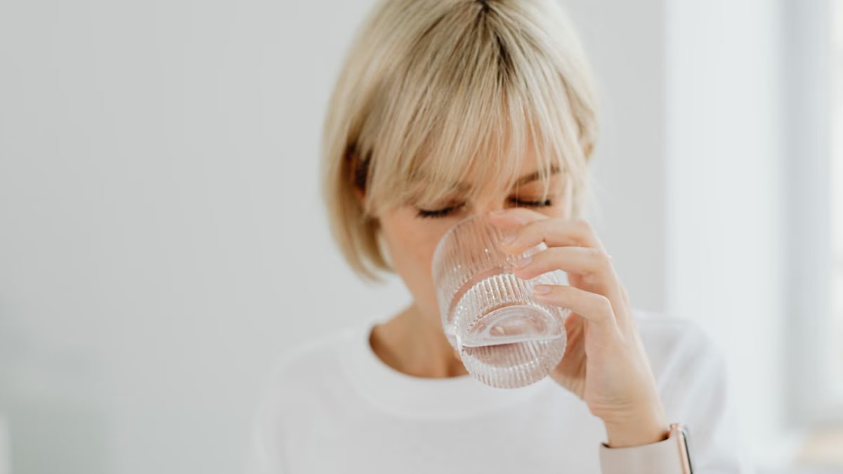 Blonde woman drinking water from glass indoors, showcasing healthy lifestyle and hydration.